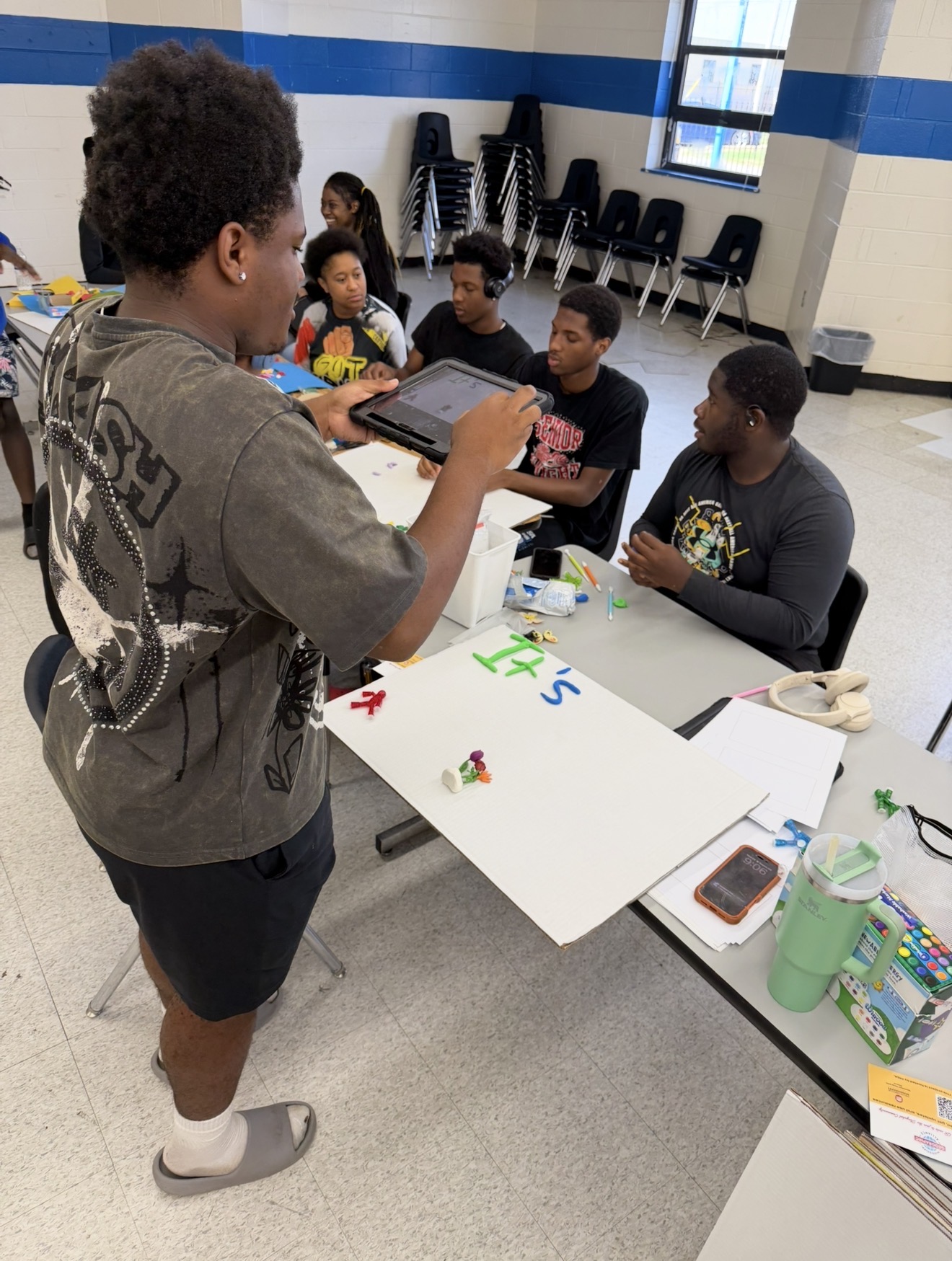 A student uses a tablet to photograph a stop-motion animation scene while classmates collaborate around a table with poster boards, molding clay, and craft materials in a classroom.