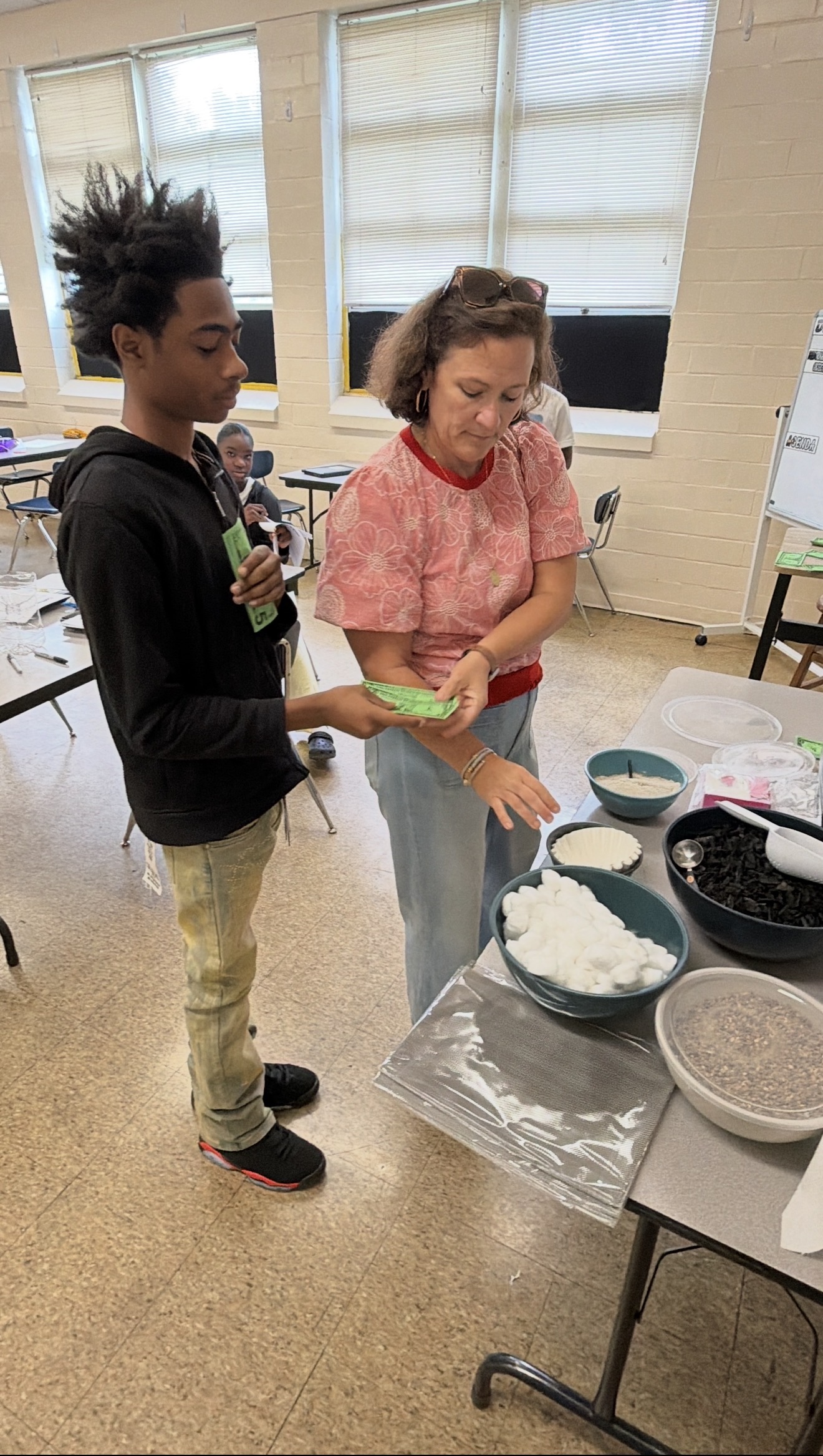 A teacher demonstrates a hands-on classroom activity to a student standing beside a table filled with materials, including cotton balls, charcoal, seeds, coffee filters, and other supplies. Other students are seated and working in the background of the classroom.