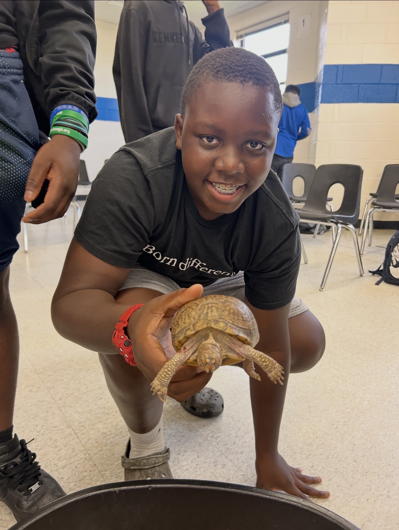 A student kneels on the classroom floor and smiles while gently holding a turtle toward the camera during a hands-on learning activity. Other students and chairs are visible in the background.
