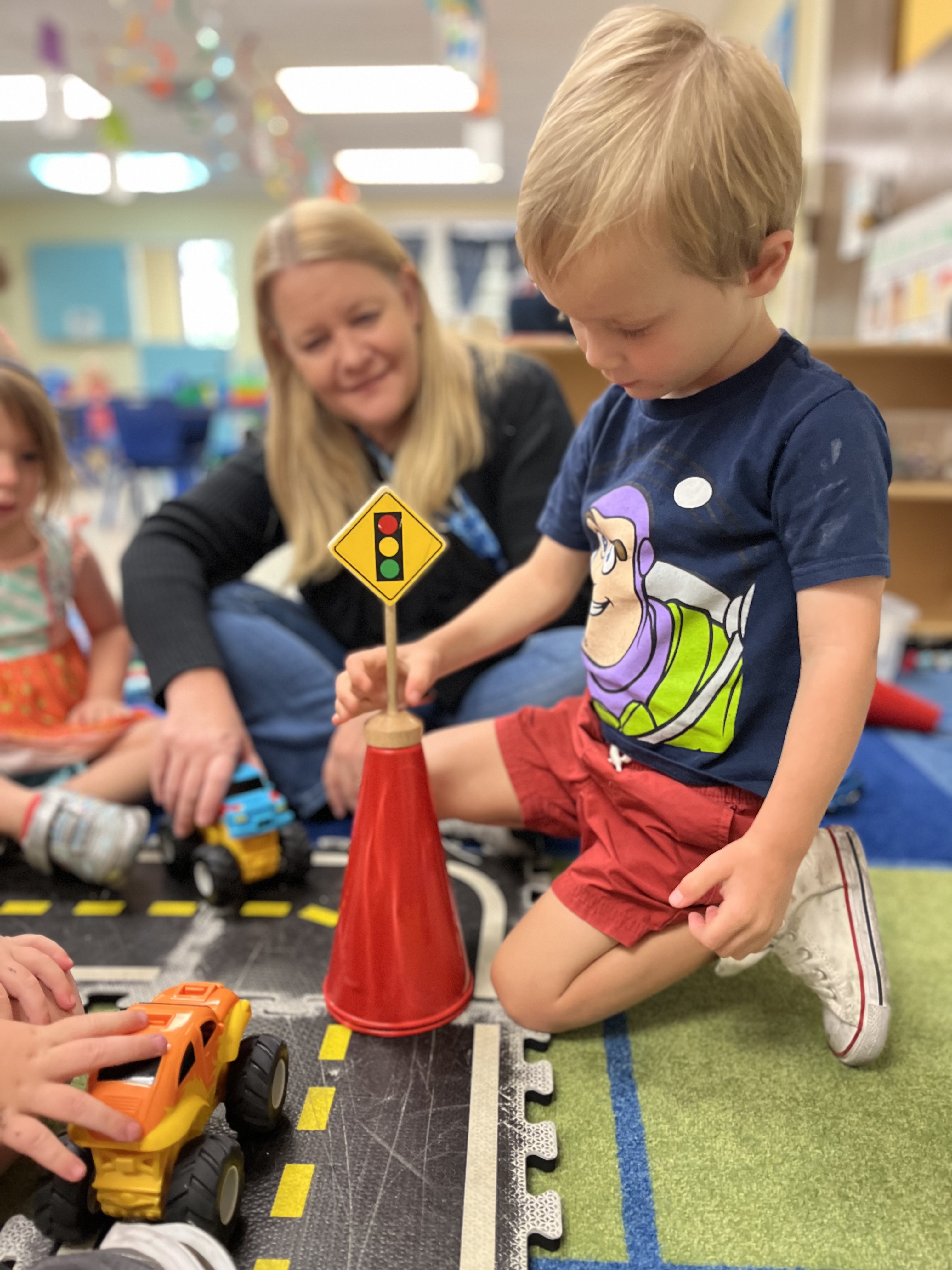A teacher watches as a young student uses wooden blocks to build a road for toy trucks.