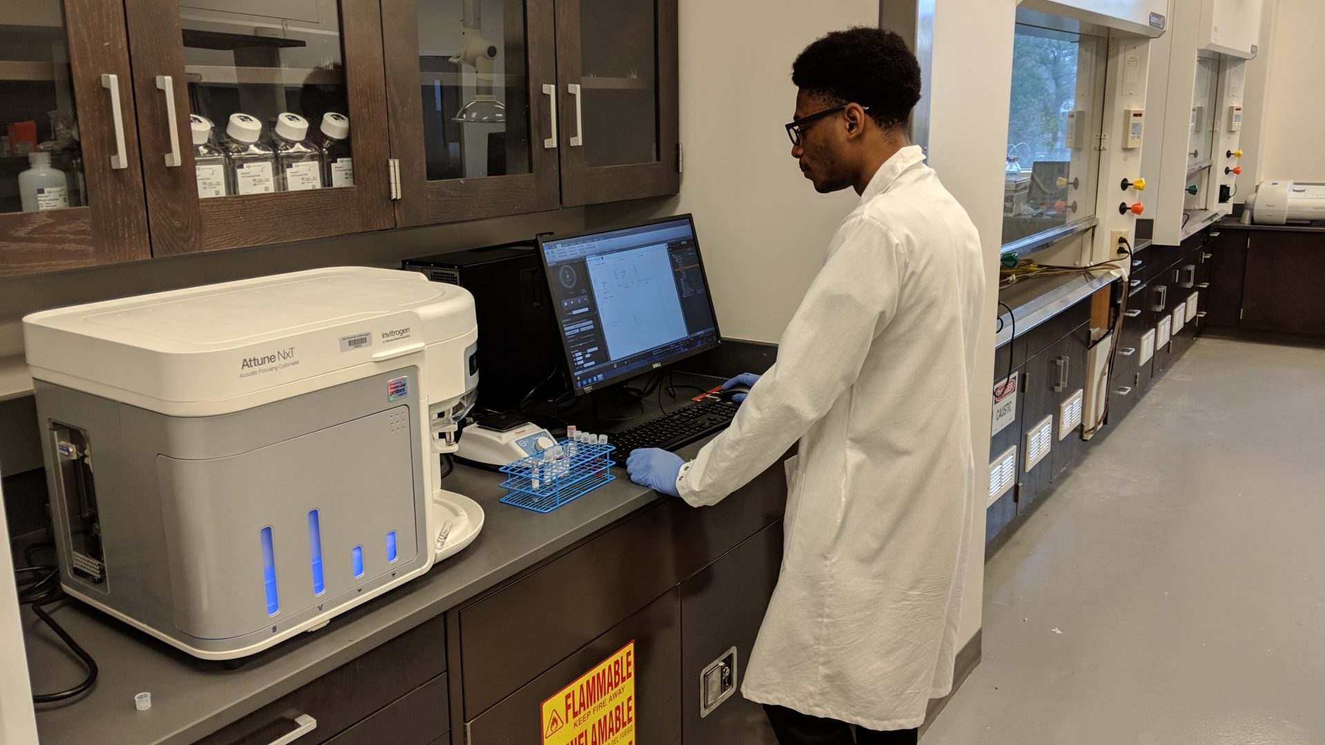 A biomedical student dressed in lab gear stands at a computer.