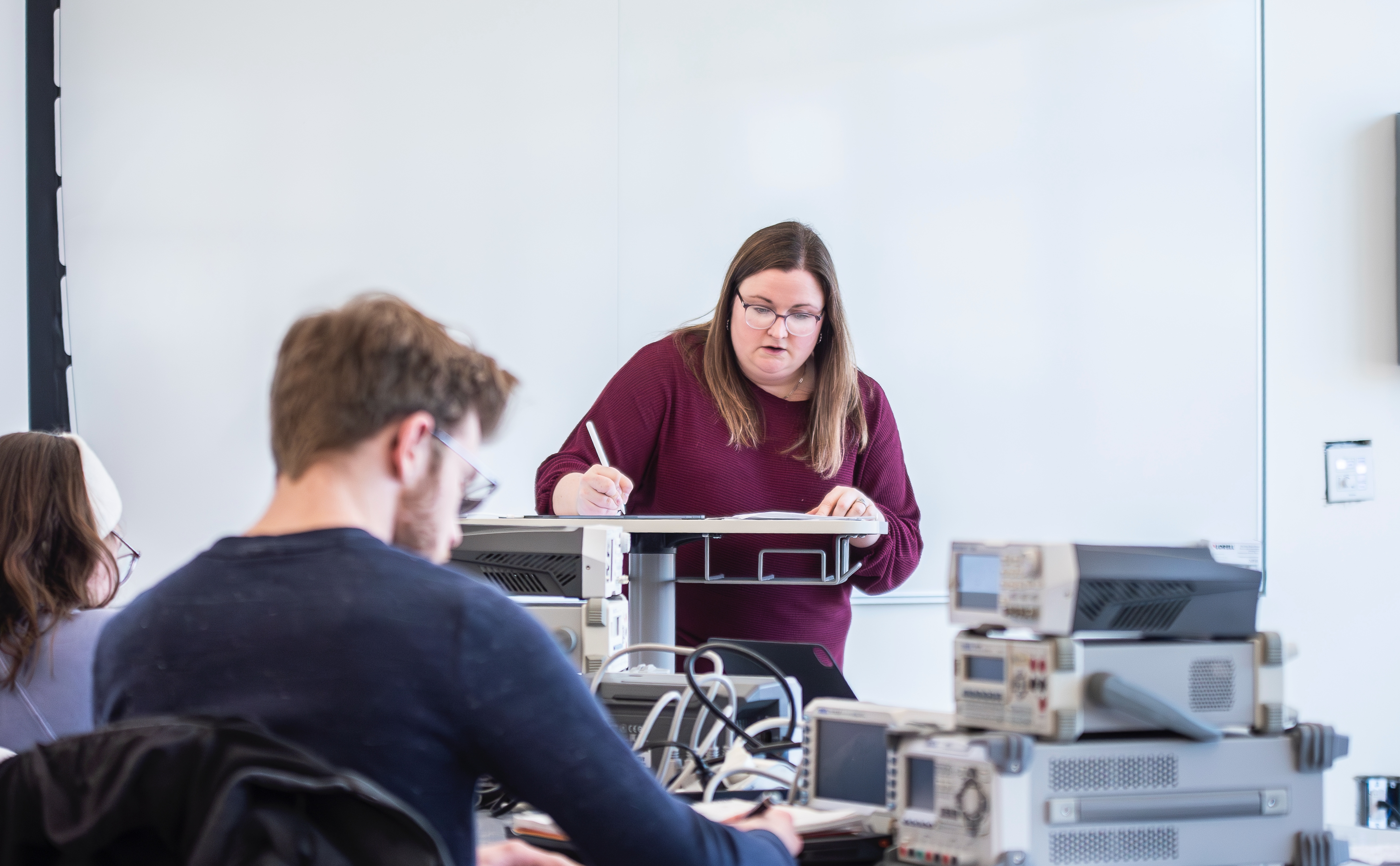 A biomedical professor stands at the front of a classroom presenting a lecture while a student takes notes.