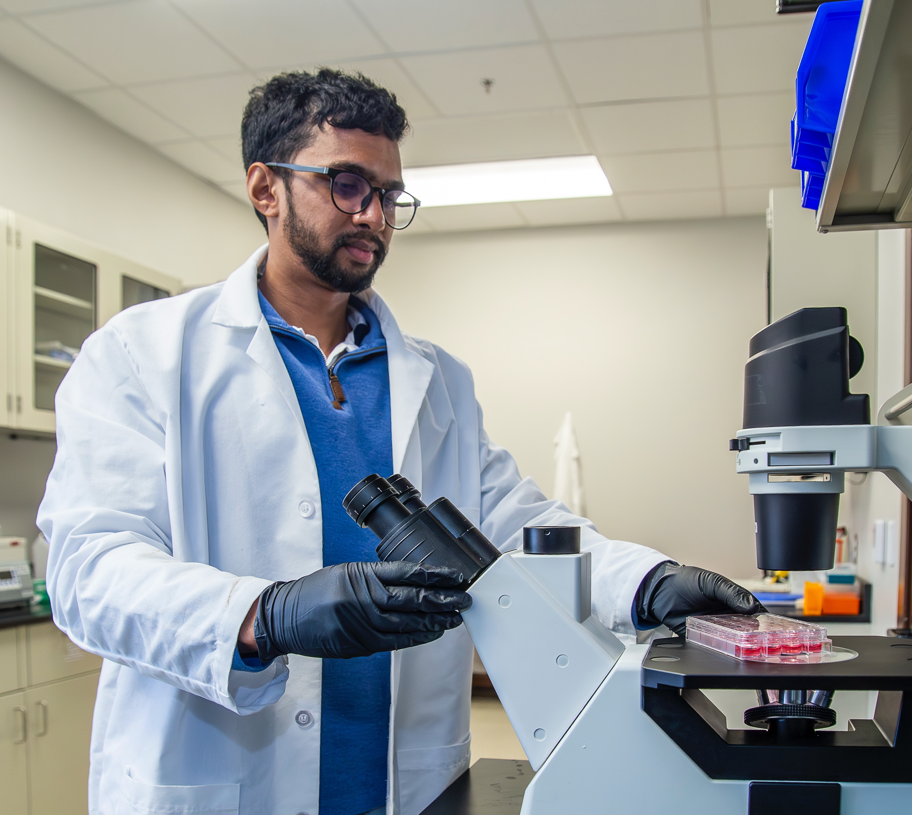 A graduate student dressed in lab equipment placing a slide on the bed of a microscope.