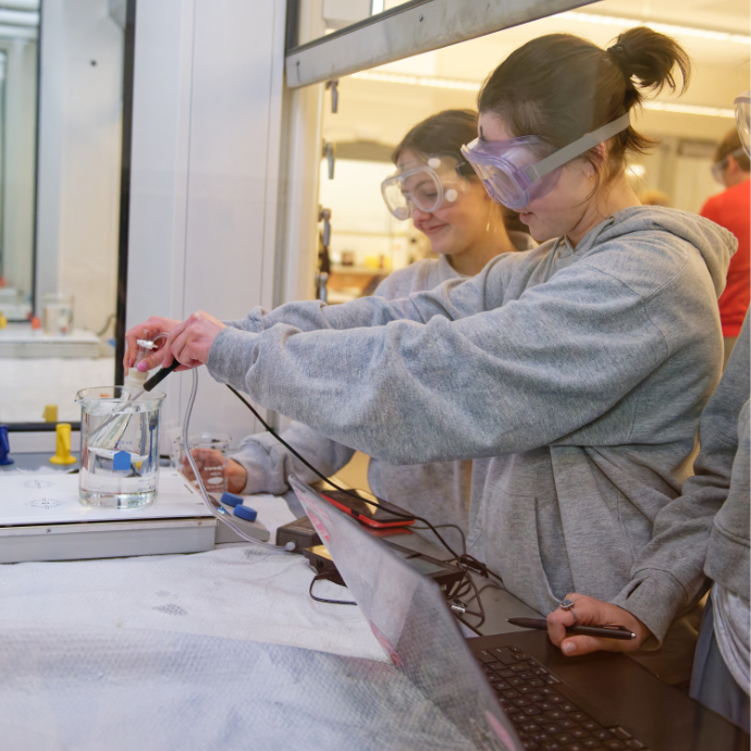 A student with safety goggles fills vials with a pipette in a laboratory with equipment in the background.