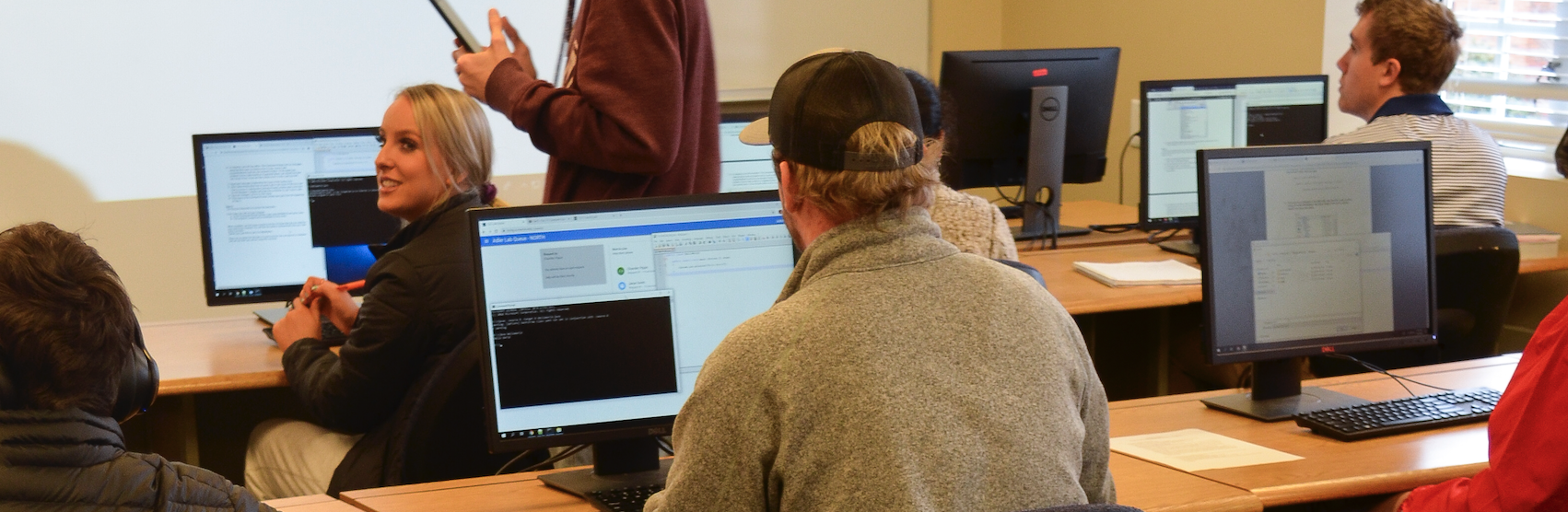 A student sits in a computer lab, surrounded by other students. He is looking at the screen where he has a command prompt open.