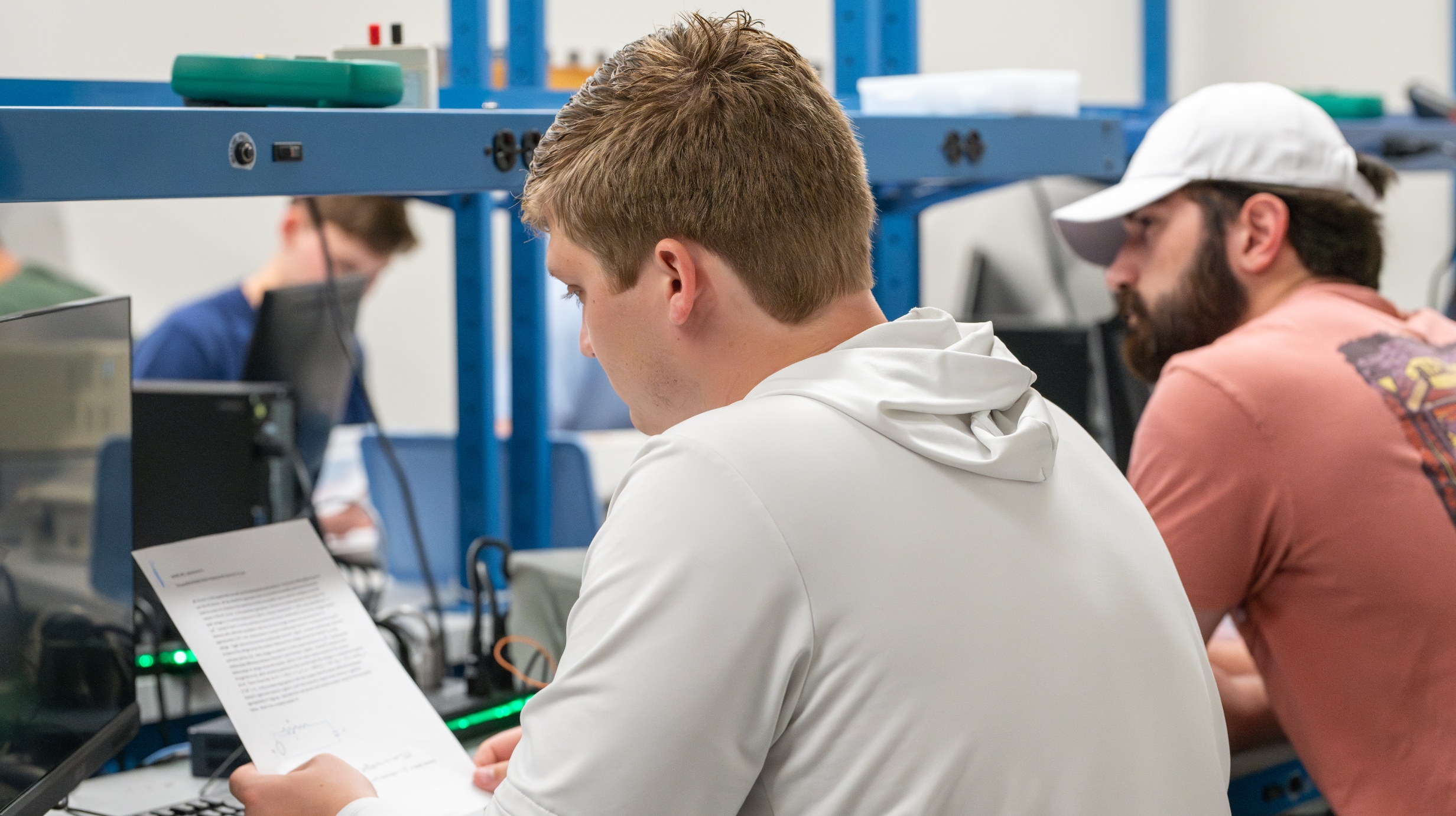 A student reviews a handout sitting at a workbench in an electrical engineering lab. Other students are sitting nearby examining their own handout.