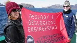 Jessee Dee and Marjorie Franks outdoors holding a banner displaying the University of Mississippi Geology and Geological Engineering logo