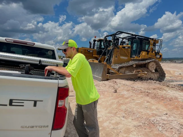 A geological engineering student rests against a truck at a work-site.  Heavy machinery can be seen in the background.