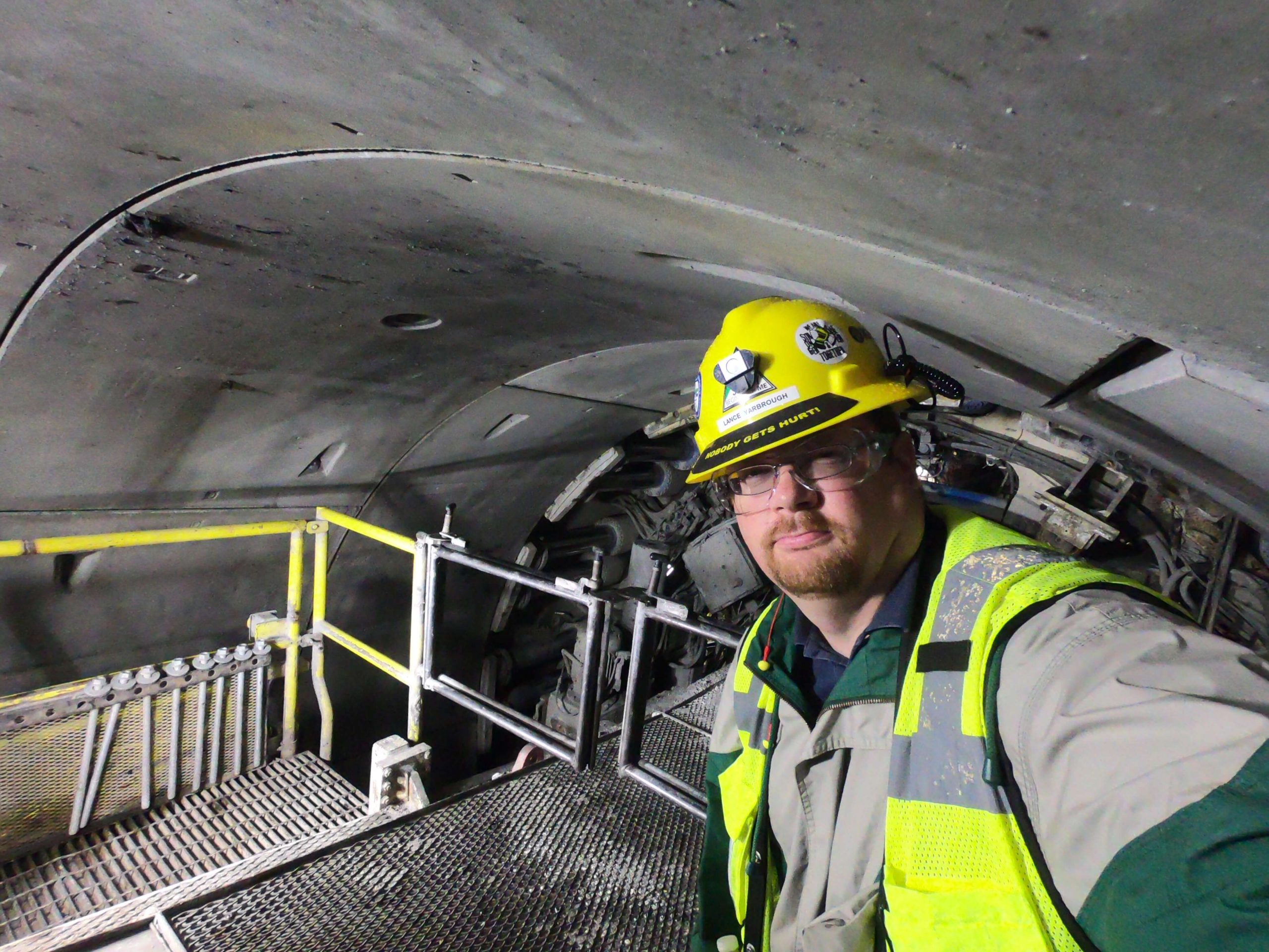 A geology professor poses for a photo in high-vis and a protective helmet next to heavy machinery. The photo appears to be underground or in a tunnel.