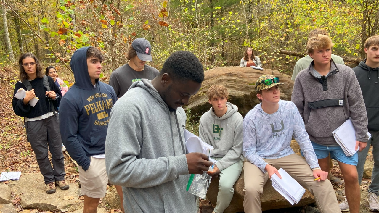 A group of students with notebooks gather outdoors look out-of-frame