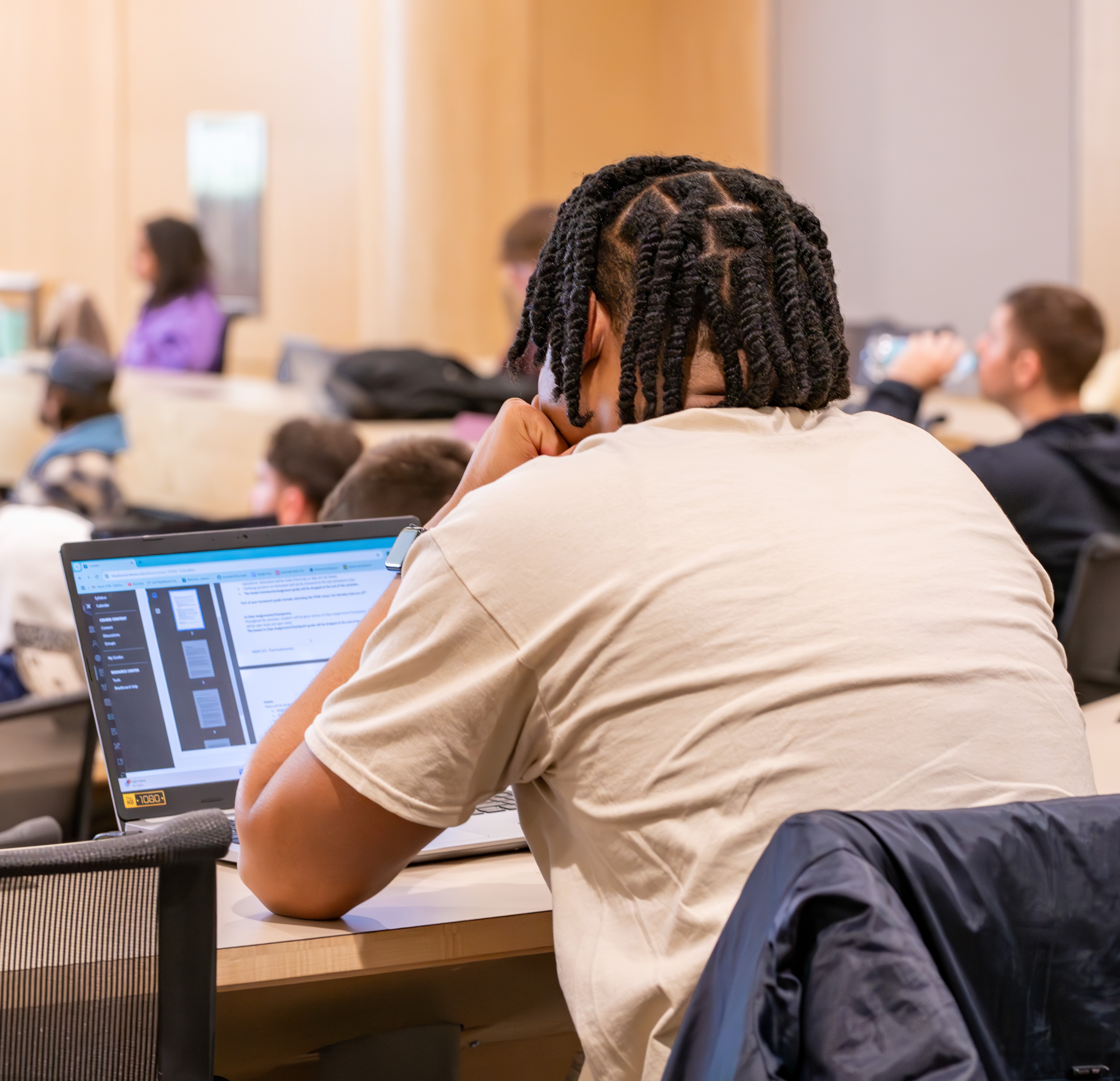 A student sits at the back of a filled classroom using a laptop.