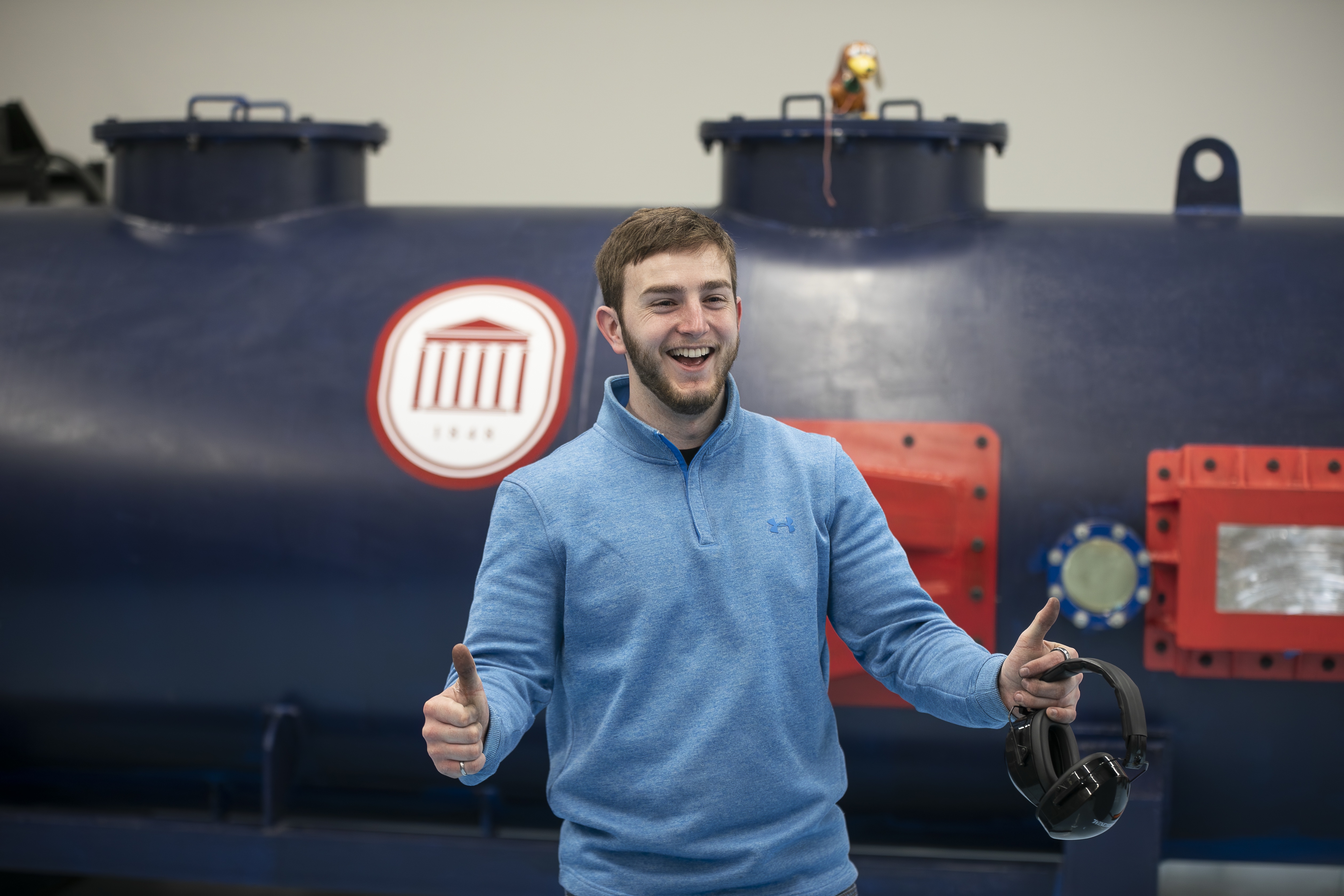 A mechanical engineering student smiles and gives two thumbs up; heavy machinery from Ole Miss's blast lab is out of focus in the background