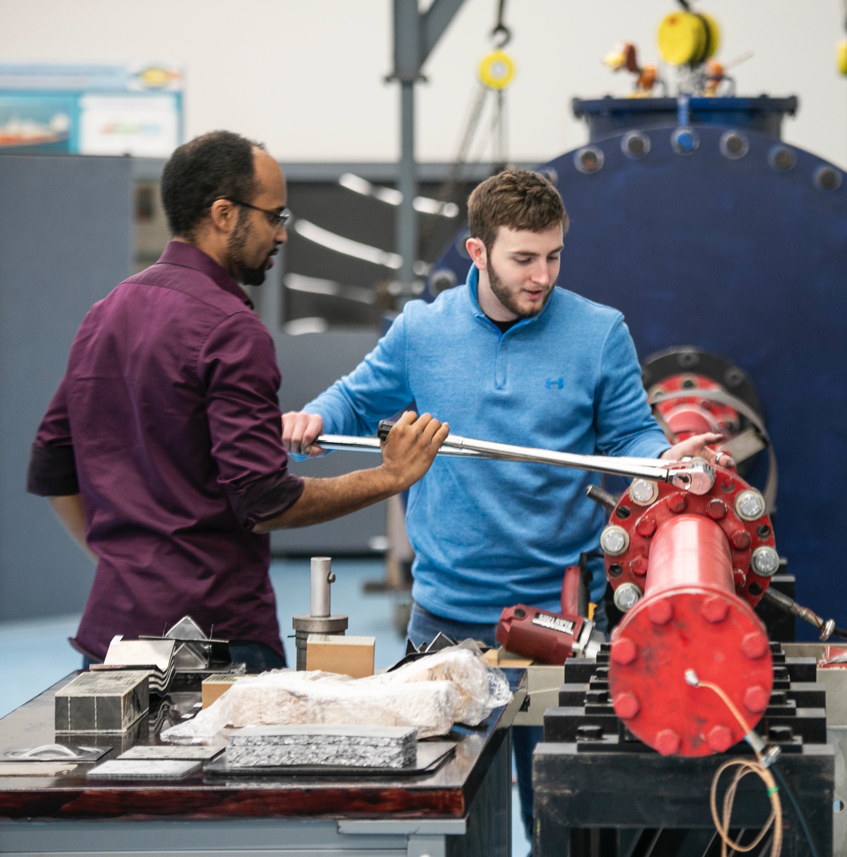 A student uses a wrench to adjust a piece of heavy machinery while an instructor provides guidance.