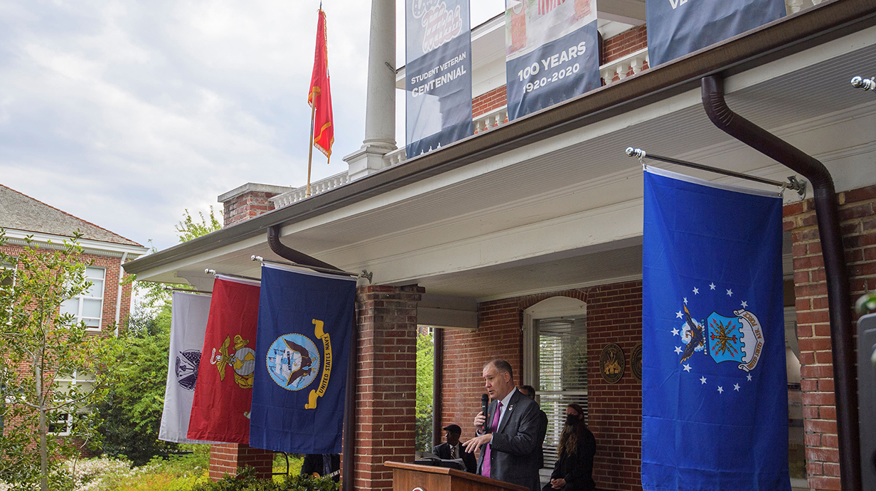 A man speaks into a microphone on a covered porch lined with flags.