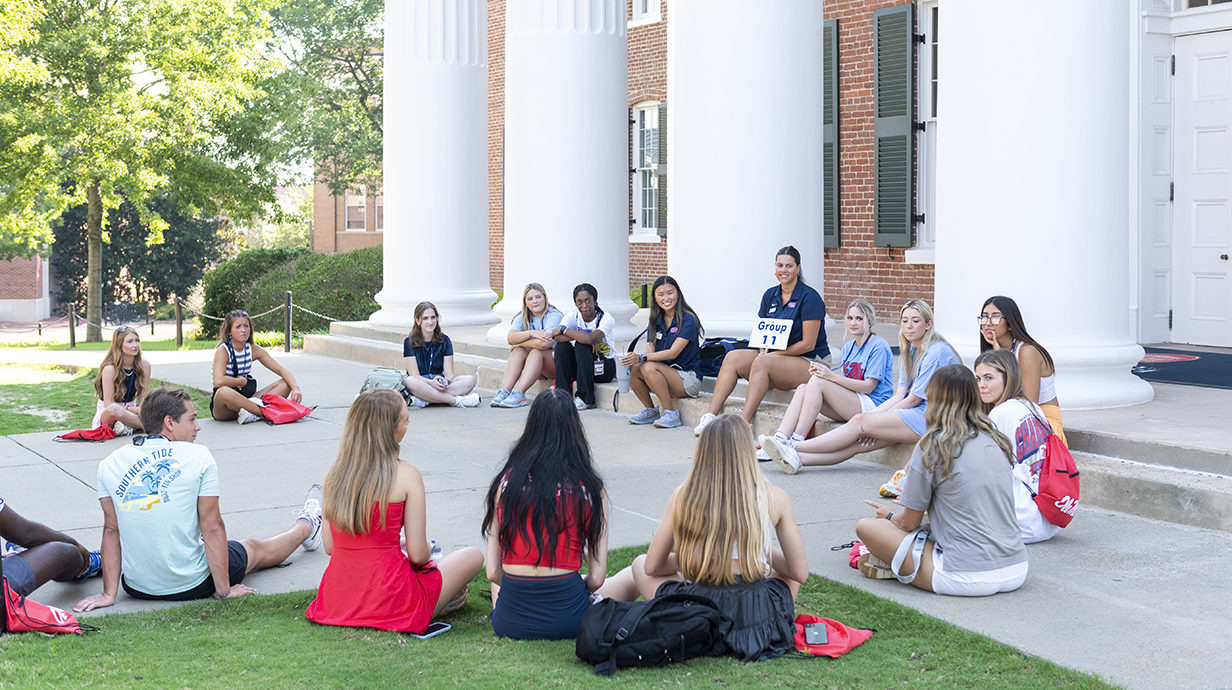 Students sit in a circle in front of the Lyceum with two Orientation Leaders during a freshman orientation session.