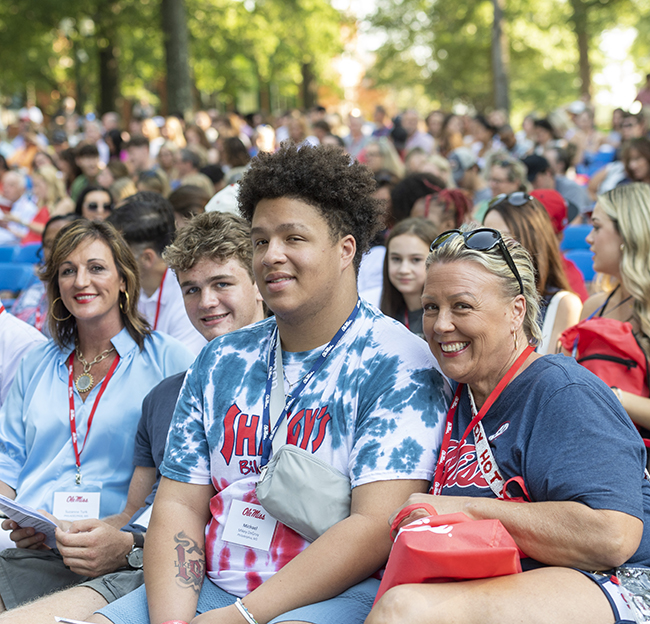 two students and their mothers sit together in the grove during orientation and smile at the camera