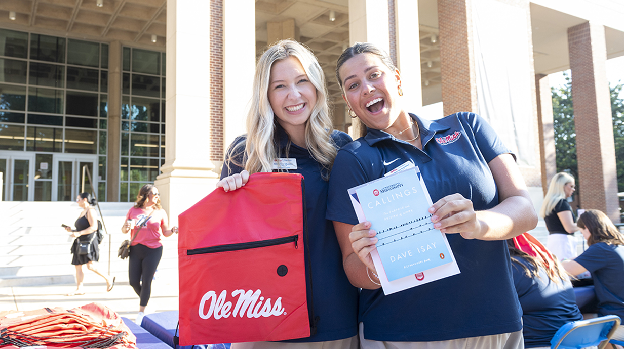 Two orientation leaders smile on Union Plaza and hold up the bag and book given to students during orientation.