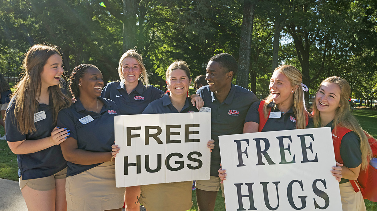 Orientation leaders hug and smile at each other in the Grove while holding two "Free Hugs" signs.