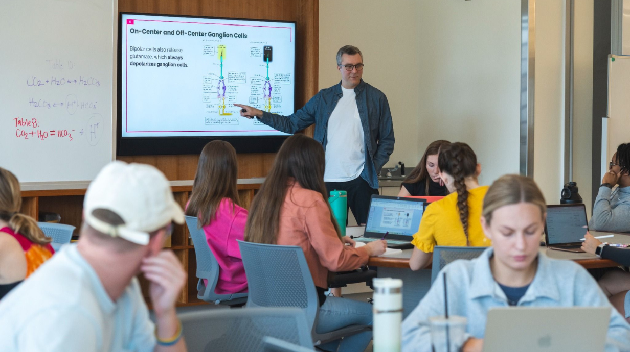 An instructor teaches in front of a projector screen with a PowerPoint slide.