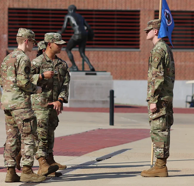 People in camoflage standing on concrete plaza. One person holds a flag