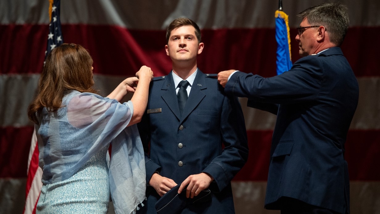 image of cadet being commissioned on a stage with parents pinning rank insignia to his coat shoulders