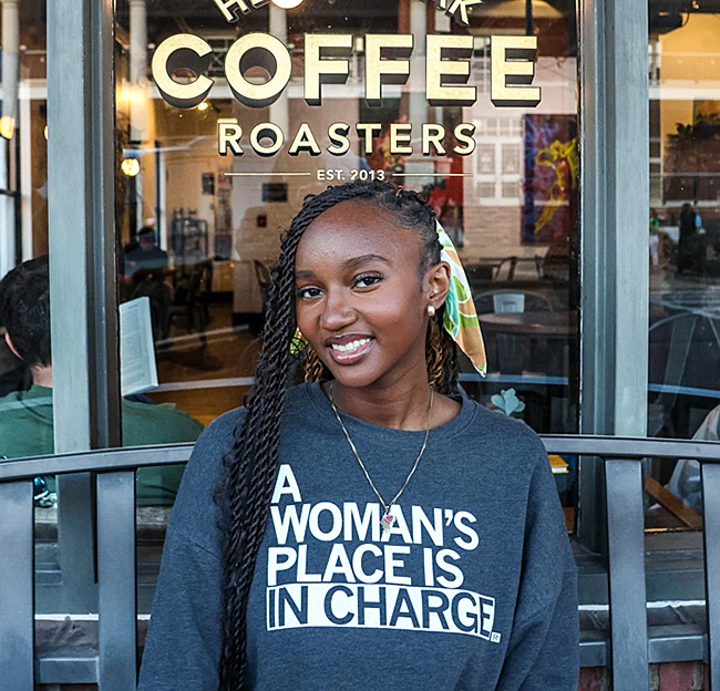 Woman wearing a sweatshirt that reads, "A woman's place is in charge," sits on bench smiling at camera; sign on window behind her reads, "Coffee"