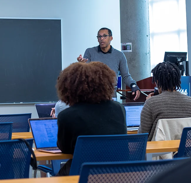 Background focus: Professor standing in front of class lecturing. Backs of students in foreground.
