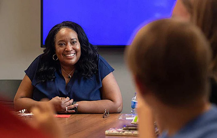 Professor sitting at table with bright blue screen behind them; out of focus students in foreground