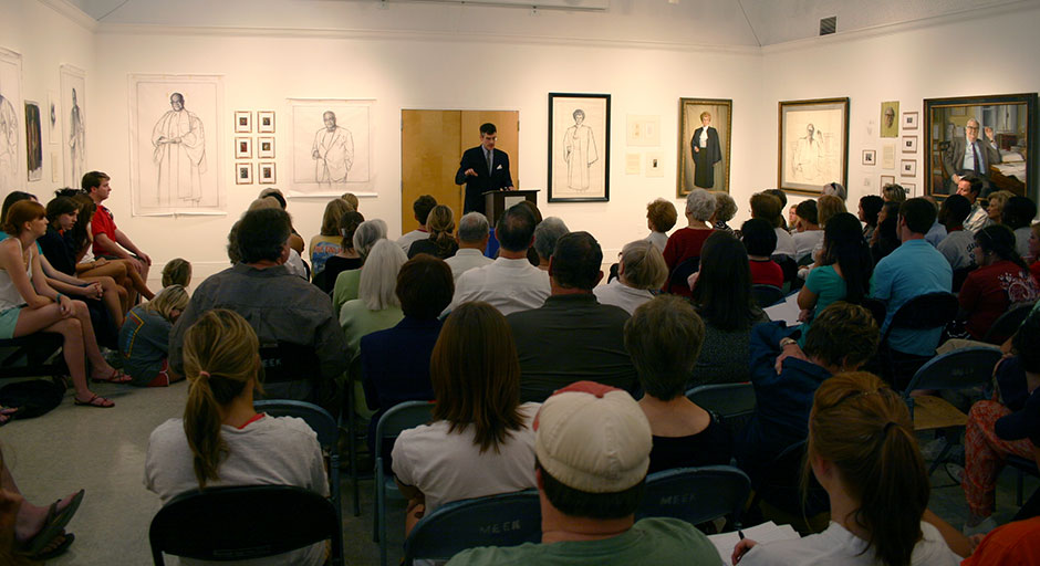 Man speaking in front of group of people discussing paintings in a gallery setting.