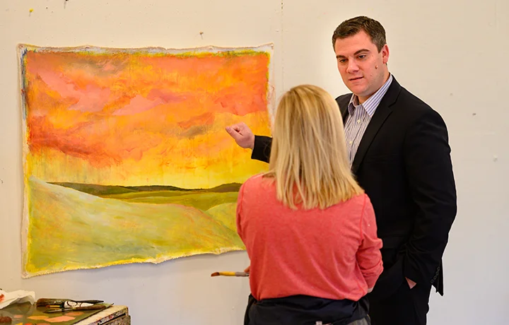 Two people stand near painting on a easel, one person is facing the camera gesturing at the painting and is facing the other person in conversation. 