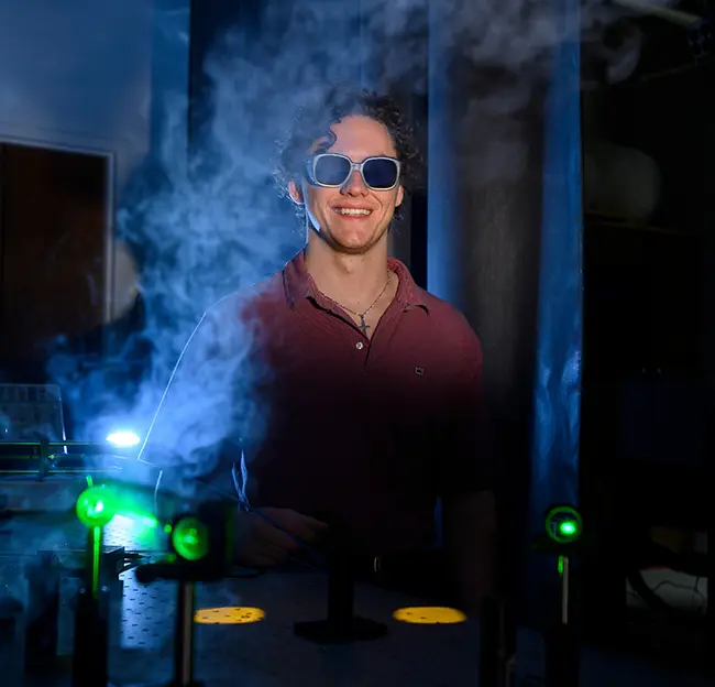 student smiling, wearing dark glasses in dark chemistry lab with blue smoke and green lights.