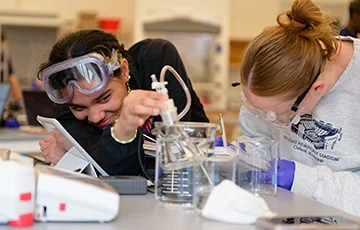 two students in lab wearing safety goggles, looking happy
