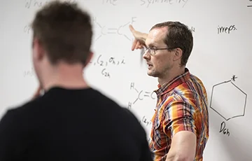 Person standing at white board with chemical diagrams written on it. Person in foreground is blurry.