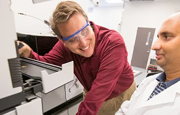 Joshua Sharp wearing safety glasses using lab equipment, other lab worker on right