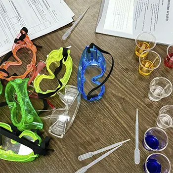 close up of goggles, papers, and beakers with colorful liquid on a table.