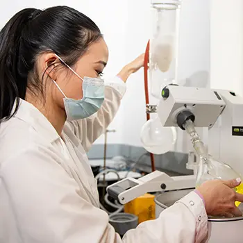 student wearing lab coat and mask works in chemistry lab; in profile; hands holding lab equipment.