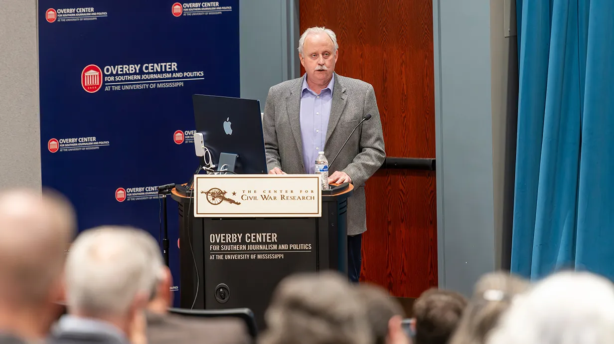 Joseph Glatthaar at podium on stage. "Overby Center for Southern Journalism and Politics at the University of Mississippi" on wall and "The center for civil war research" on a sign in front of podium.