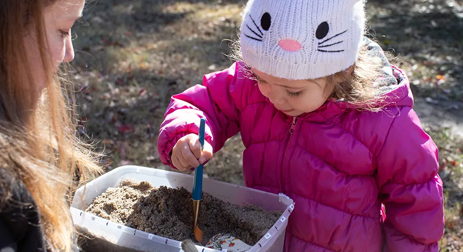 On left, child with paintbrush in dirt; adult on right holding bin of dirt.