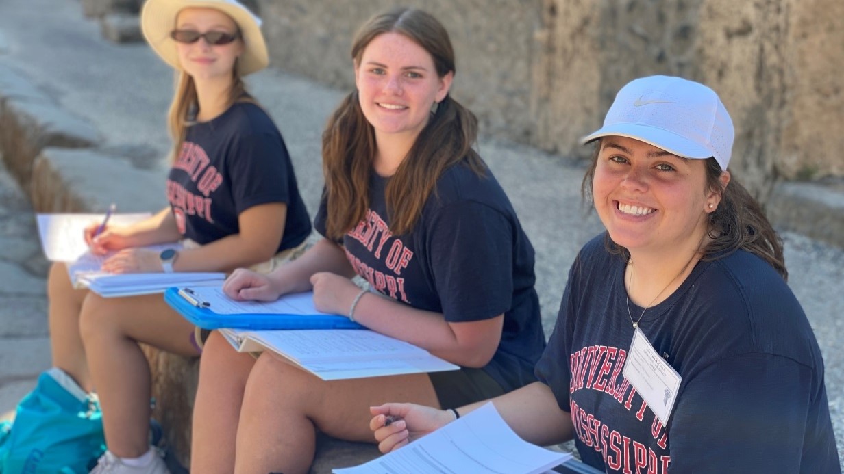 three female students seated on the ancient steps of Pompeii with clipboards and papers on their laps