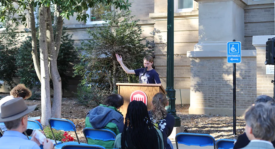 Student at a podium outdoors with arm raised addressing seated audience.