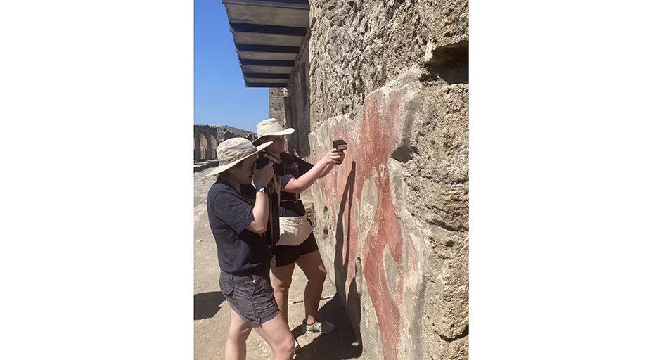 Two women standing at ancient wall. One person has a scanner in their hand, the other a camera.