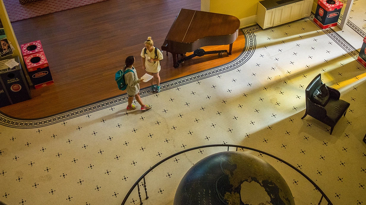 Birds eye view of entry room with black and white tiled floor, large globe of earth, piano, and two students talking.