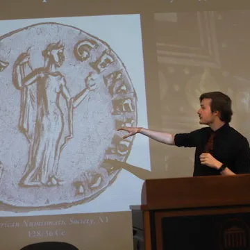 Student in front of and pointing to presentation screen. Blown up photo of ancient coin featured on screen.