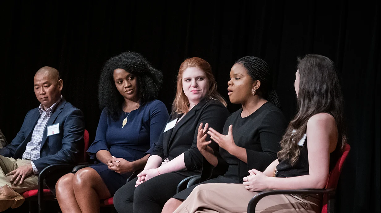 Five people wearing name tags sitting in chairs as person second to right speaks.