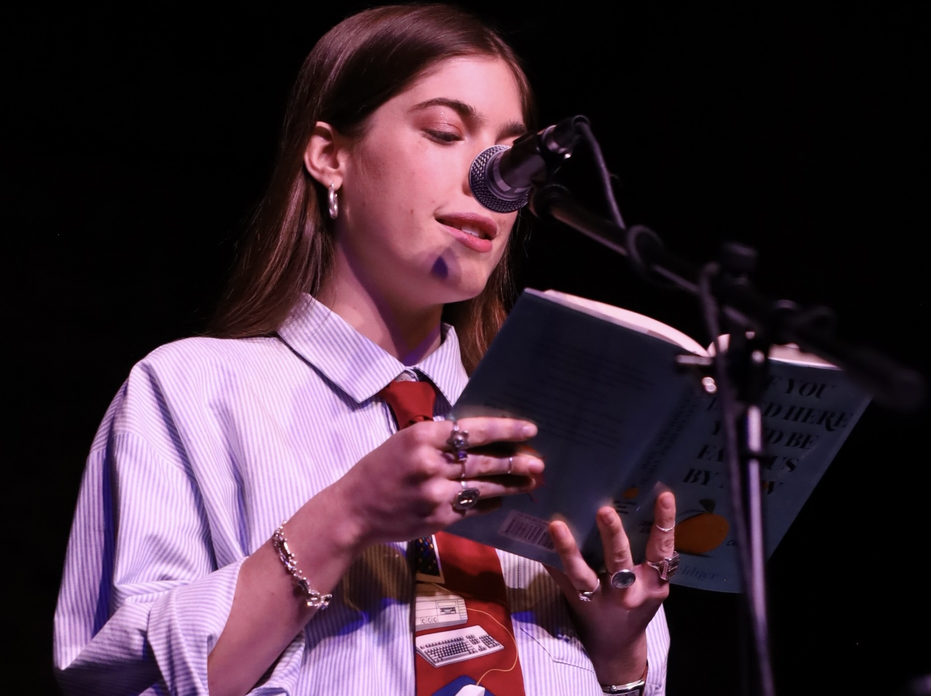 student via bleidner reads her book "if you lived here you'd be famous by now" on a darkened stage lit by blue and purple lighting