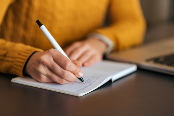 image of arms of a young person in an orange sweater as they are seated at a table while writing in a journal