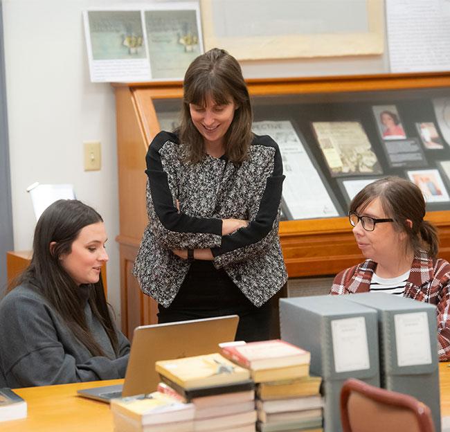 Two graduate students sit and speak to a professor standing behind them.