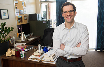 male faculty leaning on desk and smiling at camera