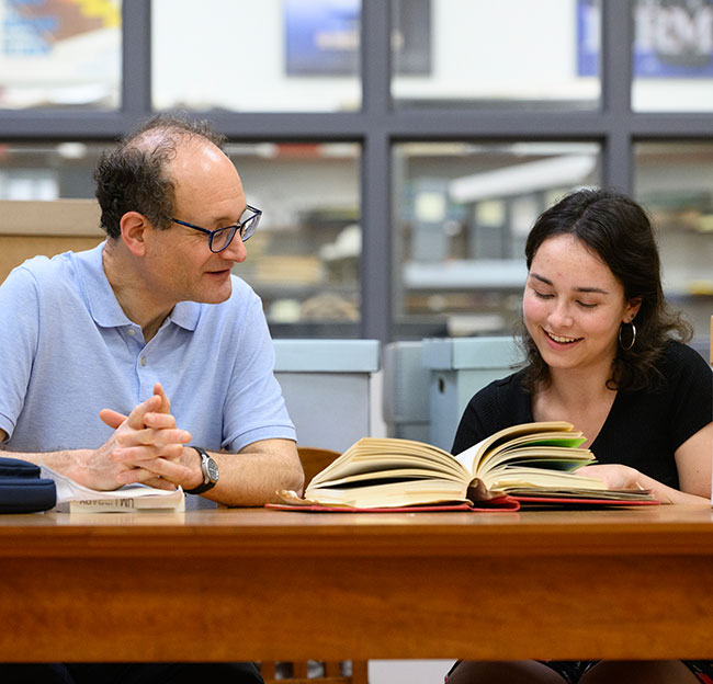 student and professor sit at desk and look through a book