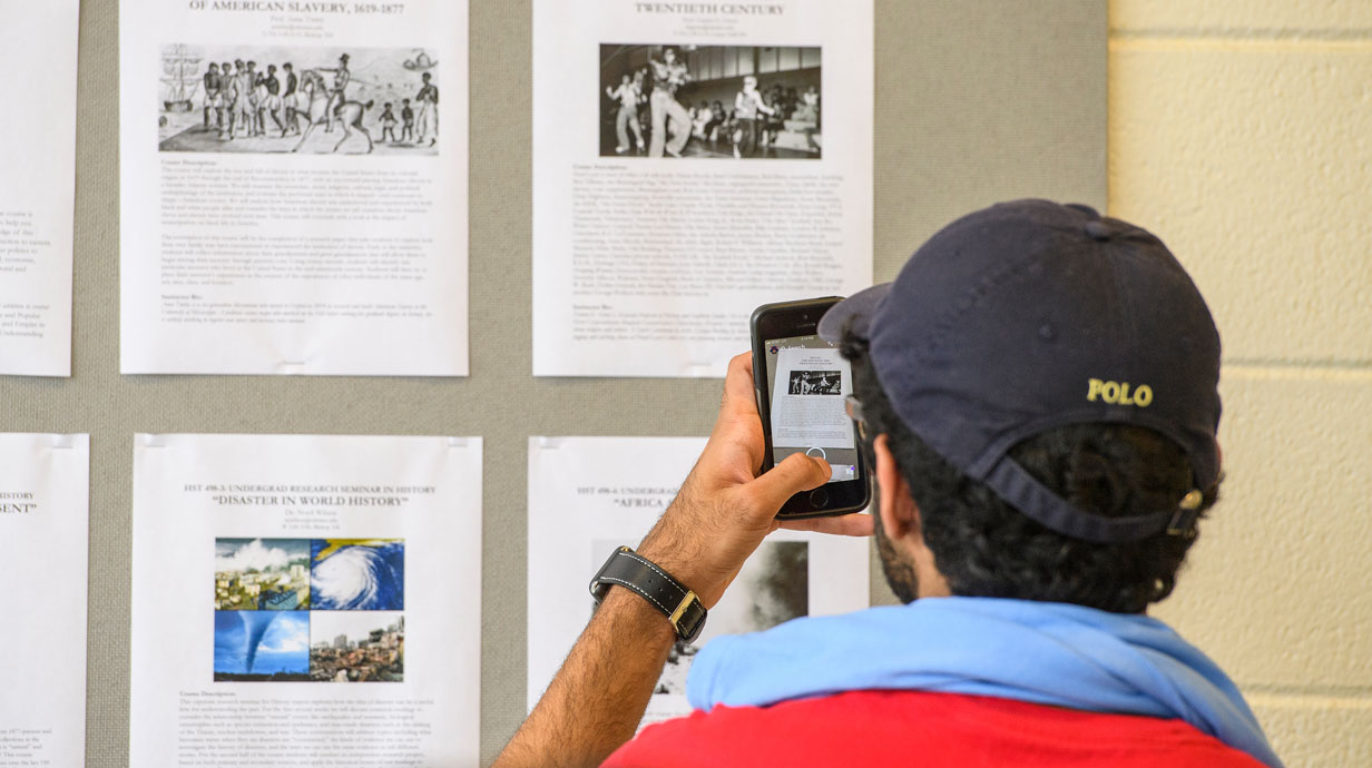 student in baseball cap taking an image of history materials hanging on the wall